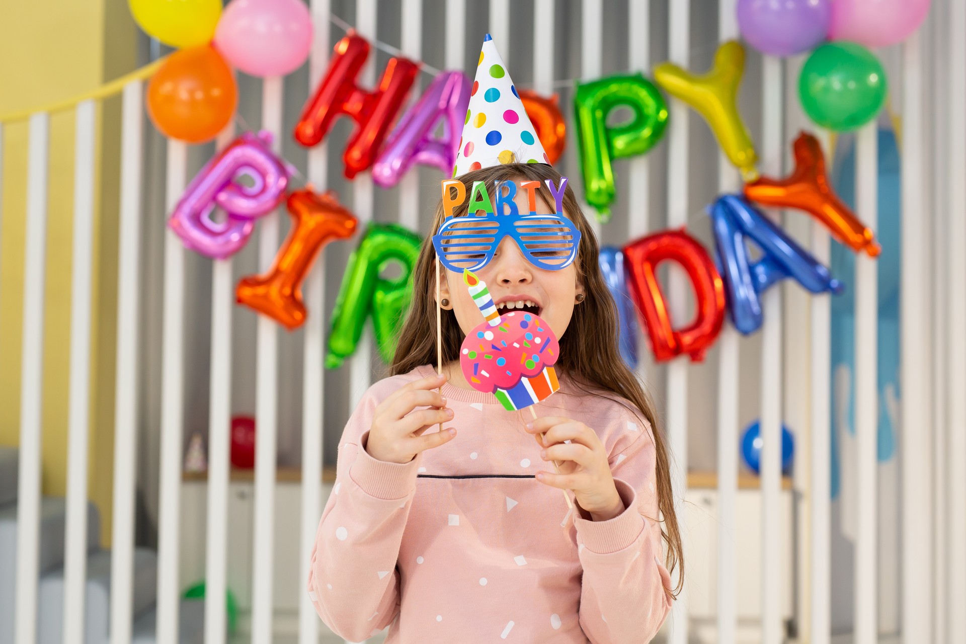 The birthday girl in a festive hat, with a pipe and props is having fun on the background of a multi-colored wall with balloons.